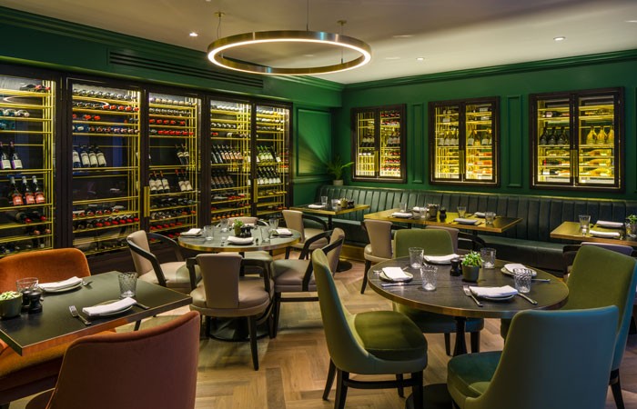 Web-The-moody,-green-wine-display-room-with-shallow-brushed-brass-chandeliers-for-added-glamour